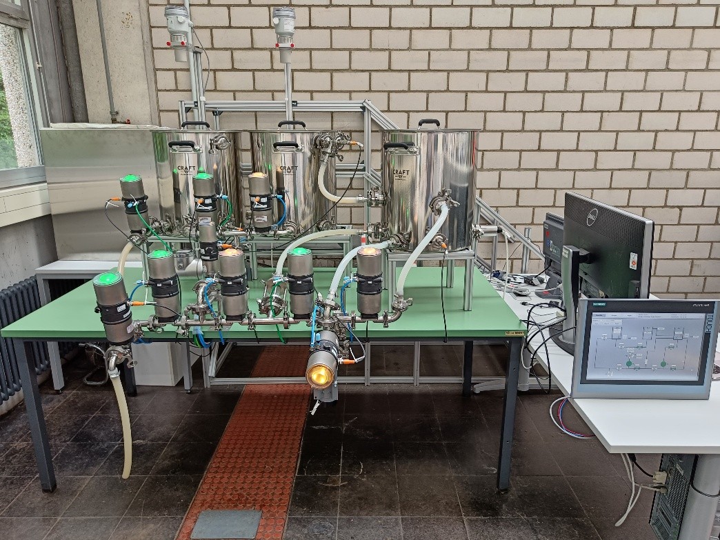 Experimentelle Brauanlage mit pneumatisch gesteuerten Ventilen, Ultraschallsensoren zur Füllstandsmessung und SPS-Steuerung; Touchscreen nicht abgebildet.